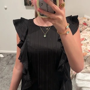 Svart blus med volangdetaljer - Säljer en svart blus med volangdetaljer längs axlar och sidor. Blusen är ärmlös och har en lätt glansig finish med diskreta vertikala ränder.