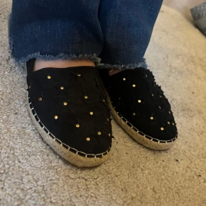 Svarta espadrillos med nitar - Säljer ett par svarta espadrillos med dekorativa guldnitar och flätad sula. Skorna har rund tå och är perfekta för vår och sommar. Snygga detaljer och bekväm passform.