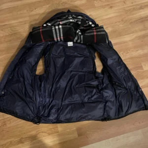 Mörkblå dunväst från Burberry - Snygg mörkblå dunväst från Burberry med klassiskt rutigt foder i huvan. Västen har dragkedja och knappar framtill samt två fickor med dragkedja. Perfekt för lager-på-lager och har en riktigt lyxig känsla.