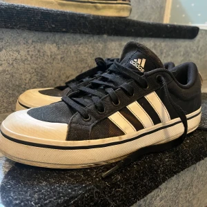 Svarta Adidas sneakers med vita ränder - Klassiska svarta sneakers från Adidas med de ikoniska tre vita ränderna på sidan och vit tå. Skorna har snörning och en platt sula, perfekt för en avslappnad streetstyle-look. Materialet är textil och syntet.