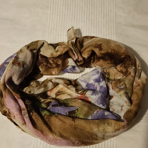 Blommig halsduk i mjukt material - Säljer en tunn och mjuk halsduk med blommigt mönster i lila, beige, brunt och inslag av rött. Perfekt accessoar för att liva upp din outfit. Halsduken är rektangulär och har en lätt glansig yta.