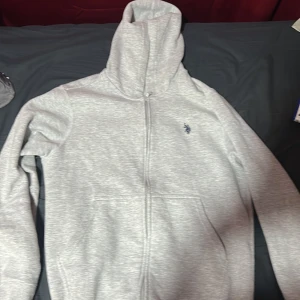 Ljusgrå hoodie med dragkedja från U.S. Polo Assn. - Säljer en ljusgrå hoodie från U.S. Polo Assn. med dragkedja och huva. Tröjan har en liten broderad logga på bröstet och är tillverkad i mjukt bomullsmaterial. Perfekt för en avslappnad stil.