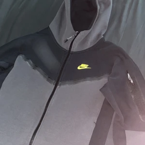 Svart och grå zip-hoodie från Nike - Nike hoodie i svart och grått med dragkedja och huva. Har en neon grön Nike-logga på bröstet och en ficka med dragkedja på ärmen. Perfekt för en sportig stil. Jätte bra skick, använt den några gånger bara. Size XS men lite större än hur XS ska sitta. 