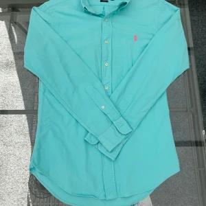 Ralph Lauren turkos skjorta – Slim Fit, stl S - Superfin Ralph Lauren skjorta i herrmodell, storlek S. Slim fit-passform som sitter snyggt utan att kännas för tight. Färgen är en riktigt fräsch turkos, och såklart med den klassiska rosa Polo-loggan broderad på bröstet.  Perfekt skjorta för våren och sommaren – funkar både till vardags med jeans eller lite mer uppklätt med chinos eller kavaj. Ett måste om du gillar preppy, stilren och tidlös look.  Skjortan är i superbra skick, använd max ett par gånger. Inga fläckar eller skador. 🔹 Storlek: S