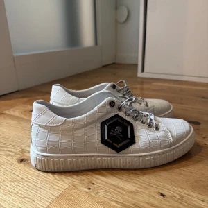 Vita sneakers från Philipp Plein - Säljer ett par vita sneakers från Philipp Plein med croco-mönstrad yta och coolt dödskallemärke på sidan. Skorna har snörning med logotyp på skosnören och präglad logga runt sulan. Perfekta för dig som vill sticka ut med stil.