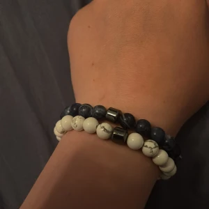 Två pärlarmband i blått och vitt från All cap one - Säljer två elastiska armband med runda pärlor. Ett armband har vita pärlor med grå marmorering och ett metallfärgat inslag, det andra har mörkblå pärlor med marmorerad effekt och liknande metallisk detalj. Perfekta att bära tillsammans eller var för sig för en stilren look.