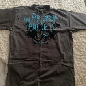 Mörkgrå t-shirt OFC Sports med tryck - Snygg mörkgrå t-shirt från OFC Sports med stort blått och svart tryck på bröstet där det står 'PACIFIC' och palmmotiv. Klassisk passform med rund hals och korta ärmar. Perfekt för dig som gillar streetstyle och grafiska prints.