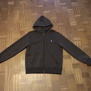 Brun Polo Ralph Lauren hoodie - Säljer en brun hoodie från Polo Ralph Lauren med dragkedja och huva. Klassisk design med två fickor framtill och den ikoniska lilla broderade loggan på bröstet. Perfekt för dig som gillar stilrena och bekväma plagg. Den är helt srillans och aldrig använd. Stl M, Passar dig som är ca 180cm lång👊🏻.                                                                         Bara och höra ev er för frågor och funderingar!👌🏻 MVH (REWEARCLOSET) Snabb affär=lägre pris!