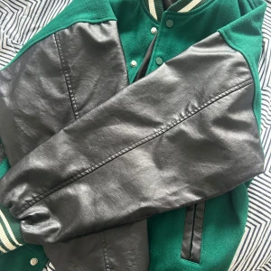 Grön och svart bomberjacka med detaljer - Snygg grön bomberjacka med svarta skinnärmar och coola broderade patches. Jackan har randiga muddar, tryckknappar och svarta detaljer vid fickorna. Perfekt för dig som vill sticka ut med en sportig och trendig look.