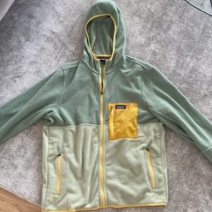 Grön fleecejacka med huva från Patagonia - Säljer en grön fleecejacka från Patagonia med huva och dragkedja. Jackan har gula detaljer, två sidofickor med dragkedja och en gul bröstficka. Perfekt för kyliga dagar och outdoor-aktiviteter. Är använd någon enstaka gång men Hoodien är i topp skick. 