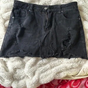 Svart jeanskjol med slitna detaljer - Svart kort jeanskjol i 100% bomull med rå kant och slitna detaljer framtill. Klassisk femficksmodell med dragkedja och knapp. Perfekt för en edgy och avslappnad stil.