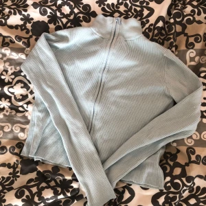 Ljusblå ribbad kofta från Wow - Säljer en ljusblå ribbad kofta från Wow med lång ärm och dragkedja framtill. Tröjan har en hög krage och är i mjukt, stickat material. Perfekt att slänga på sig när det är lite kyligare ute. Och lite kortare i magen. 