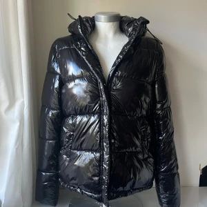 Svart glansig pufferjacka från ONLY - Säljer en supercool svart glansig pufferjacka från ONLY. Den är perfekt för kalla dagar med sin vattenavvisande och vindtäta design. Jackan har en dragkedja framtill och en mysig huva. Den är i nyskick och redo för vinterns äventyr!