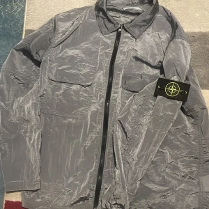 Stone Island overshirt  - Säljer en grå overshirt från Stone Island med klassisk patch på ärmen. Jackan har två bröstfickor med lock, hel dragkedja och är tillverkad i lätt, glansig syntet. Perfekt för lager-på-lager och streetwear-vibe. Unik färg Storlek M