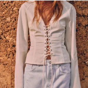 Korsett topp Zara - Supersnygg vit korsettopp med långa ärmar och djup v-ringning. Toppen har snörning framtill som ger en riktigt cool och trendig look. Figurnära passform och lätt croppad längd. Perfekt att styla med jeans eller kjol för en edgy vibe.