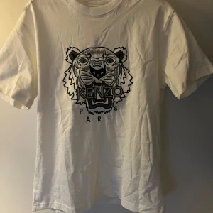 Vit Kenzo t-shirt med tigertryck - Vit t-shirt från Kenzo med ikoniskt svart tigertryck och texten 'KENZO PARIS' framtill. Klassisk passform med rund hals och korta ärmar. Tillverkad i mjuk bomull som känns skön mot huden. Perfekt för dig som vill sticka ut med en stilren och trendig look.