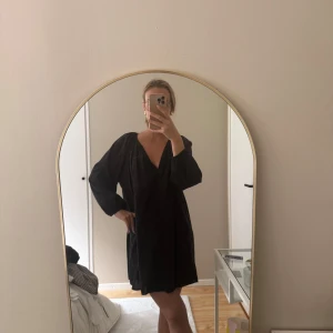 Svart kortklänning med v-ringning - Säljer en svart kortklänning med v-ringning och långa, lösa ärmar. Klänningen har en avslappnad passform och är perfekt för dig som gillar en enkel men stilren look.