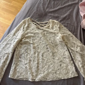 Vit spetsblus med korsade band - Säljer en vit blus i spets med stora blommönster och långa ärmar. Blusen har v-ringning med dekorativa korsade band framtill. 