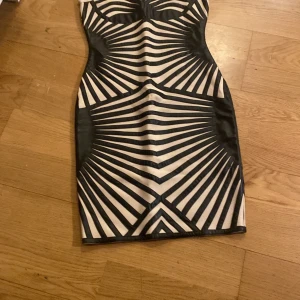Svart och beige tight klänning med grafiskt mönster - Säljer en tight klänning i svart och beige med ett grafiskt, randigt mönster. Klänningen är axelbandslös och har dragkedja bak. Perfekt för dig som vill sticka ut med en unik look.