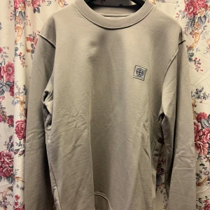  Grå sweatshirt från Stone Island - Säljer en grå sweatshirt från Stone Island med rund hals och diskret logotyp på bröstet. Tröjan har långa ärmar och är tillverkad i mjuk bomull, perfekt för en stilren och avslappnad look.
