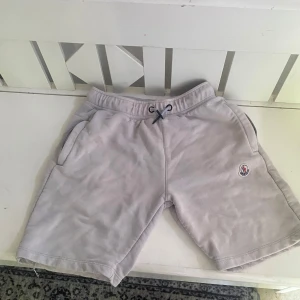 Ljusgrå Moncler shorts med snörning - Snygga ljusgrå shorts från Moncler med klassisk logga på ena benet. Modellen har elastisk midja med svart snörning och två sidofickor. Mjukt material som känns skönt mot huden, perfekt för sommaren eller när du vill ha en clean look.