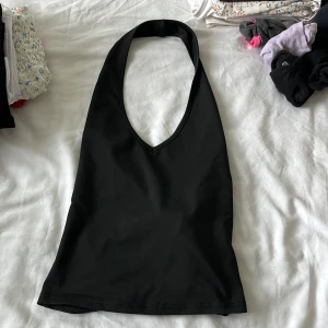 Svart halterneck linne - Snygg svart halterneck linne med djup v-ringning och breda axelband som knäpps i nacken. Toppens stilrena design gör den enkel att matcha med olika outfits. Materialet känns mjukt och stretchigt, perfekt för varma dagar.