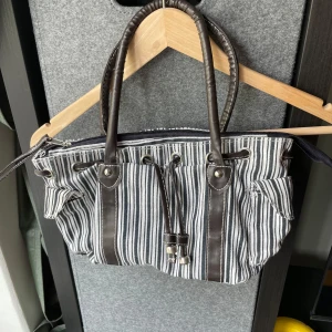 Randig axelväska secondhand - Säljer en snygg axelväska med bruna ränder och bruna skinnhandtag. Väskan har dragkedja upptill, två sidofickor och dekorativ snörning framtill. Perfekt för dig som gillar en stilren men ändå unik look.