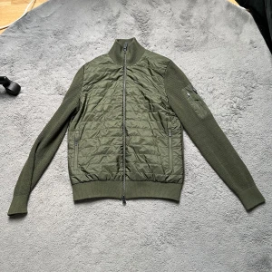 Olivgrön hybridjacka från J.Lindeberg - Snygg olivgrön jacka från J.Lindeberg med quiltad front och rygg i stickat material. Jackan har dragkedja framtill, ribbade muddar och en ärmficka med logotyp på vänster ärm. Perfekt för lager-på-lager och stilren look.