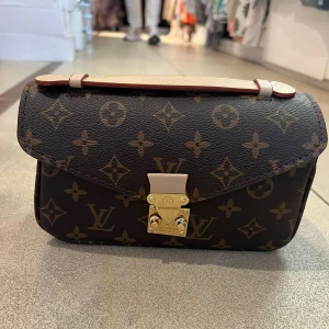 Anitas monogram axelväska - Säljer en klassisk axelväska  med ikoniskt monogrammönster i brunt och beige. Väskan har guldfärgat spänne och ett ljust handtag upptill. Perfekt för dig som vill ha en tidlös accessoar med lyxig känsla.