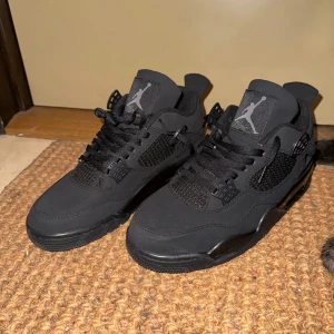 Svarta Nike Air Jordans 4 - Säljer ett par oanvända helsvarta Nike Air Jordans 4 med klassisk siluett och Jumpman-logga på plös och häl. Skorna har snörning, meshpaneler på sidorna och en platt sula. Perfekta för dig som gillar streetwear och vill ha en stilren look.