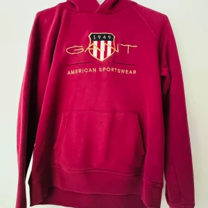 Röd hoodie från Gant - Säljer en röd hoodie från Gant med broderad logga och texten 'American Sportswear' framtill. Tröjan har huva och en stor magficka. Perfekt för dig som gillar klassisk och sportig stil.