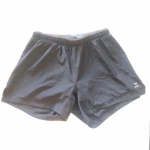 Svarta träningsshorts från SOC - Svarta träningsshorts från SOC med elastisk midja och kort passform. Perfekta för löpning eller gym, tillverkade i lätt syntetmaterial som andas. Diskret logga på ena benet och klassisk sportig look.