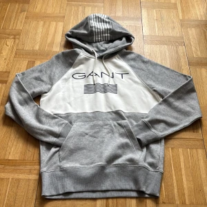 Grå och vit hoodie från GANT - Snygg hoodie från GANT i grått och vitt med stor logga framtill och vågigt tryck. Tröjan har huva med snörning och coola ränder på luvan. Mjuk bomullsmix, känguruficka och långa ärmar. Perfekt för chill dagar eller när du vill ha en clean look.