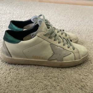 Golden Goose Superstar sneakers med grön häl - Säljer ett par vita Golden Goose Superstar sneakers med grå stjärndetalj på sidan och grönt parti vid hälen. Skorna har snörning och platt sula, samt en klassisk rund tå. Perfekta för dig som gillar en stilren men ändå unik look. Knappt använda! Priset kan diskuteras! ‼️obs har en defekt som man ser på fjärde bilden‼️