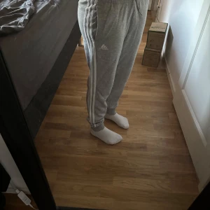 Grå mjukisbyxor från Adidas - Säljer ett par grå mjukisbyxor från Adidas med klassiska vita ränder längs sidorna och logga på låret. Byxorna har resår i midjan och är perfekta för en avslappnad stil.