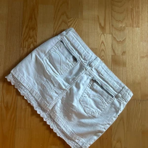 Vit jeanskjol med detaljer - Säljer en vit jeanskjol med slitna detaljer och spetskant längst ner. Kjolen har fickor med coola dragkedjor där bak. Perfekt till sommaren! 