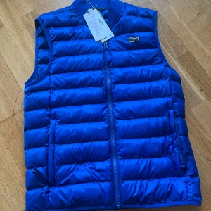 Blå dunväst från Lacoste - Mörk blå väst i märket Lacoste, det finns en bränn skada i västen som du kan se på sista bilden men är väldigt liten och syns inte vid användning eftersom det sitter på insidan, annars skick 10/10 nästan helt ny, nypris ligger på runt 1100 kr så är en bra steal, hör av er vid frågor