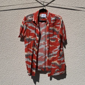 Röd mönstrad skjorta från Vaillent - Säljer en röd kortärmad skjorta från Vaillent med grått och vitt fågelmönster och tryckt text. Skjortan har klassisk krage, knappar framtill och är tillverkad i 100% viskos. Storlek S.