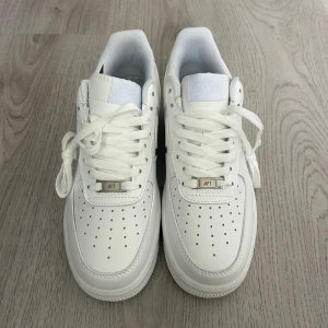 Vita Nike Air Force 1 sneakers - Helt nya klassiska helvita Nike Air Force 1 sneakers med perforerad tå, snörning och låg profil. Skorna har Nike-logga på sidan och bak, samt en platt sula för skön komfort. Perfekta för en clean och stilren look.