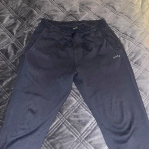 Svarta mjukisbyxor från BOSS - Svarta mjukisbyxor från BOSS med dragsko i midjan och diskret logga på vänster sida. Byxorna har en avslappnad passform och är perfekta för en chill stil. Mjukt material och elastisk midja för extra komfort. Byxorna är lite distressed men inte slitna.