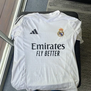 Real Madrid Mbappé matchtröja Adidas - Snygg vit Real Madrid fotbollströja med Mbappé och nummer 9 på ryggen. Tröjan är ifrån denna fotboll säsongen. Tröjan har svarta detaljer, klubbmärke på bröstet, Adidas-logga och sponsortryck. Kortärmad modell i lätt funktionsmaterial med Champions League-märke och UEFA Foundation-patch på ärmen.