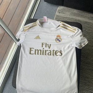 Real Madrid vit matchtröja Adidas - Snygg vit Real Madrid fotbollströja från Adidas med guldiga detaljer och klubbmärke på bröstet. Tröjan har korta ärmar, rund hals och klassiska Adidas-ränder på axlarna. Materialet är lätt och andas, perfekt för fotboll eller träning.