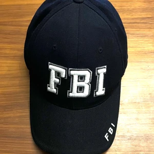  FBI- broderad keps - Svart - Snygg svart keps från Rapid Dominance med broderad FBI-text i vitt framtill och baktill. Kepsen har en justerbar rem för perfekt passform och är tillverkad i 100% akryl.