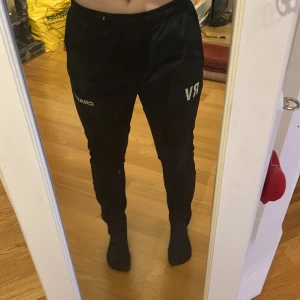 Svarta träningsbyxor med tryck - Snygga svarta träningsbyxor med elastisk midja och tryck på benen. Perfekta för en aktiv livsstil. Byxorna har en bekväm passform och är lätta att röra sig i. Det finns initialer RV från fotboll. Och även en dragkedja längst ner på båda benen för att få på sig de lättare.