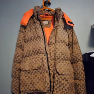 Mönstrad dunjacka från Gucci/adidas  - Säljer en snygg och varm dunjacka från Gucci/adidas med ett unikt mönster i brunt och beige. Jackan har en iögonfallande orange insida och praktiska fickor framtill. Perfekt för kyliga dagar! Har köpt begagnade men vi har aldrig använt den då den var för stor för sonen. Osäker på äkthet.