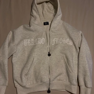 Grå hoodie från Brand of roses - Säljer en grå hoodie från Brand Roses med dragkedja och huva. Den har ett tryck i vitt på bröstet och två praktiska fickor framtill. Perfekt för en avslappnad stil.