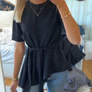 Svart blus med knytband - (Intressekoll) Snygg svart blus i kostym tyg med korta ärmar och ett knytband i midjan för en smickrande passform. Perfekt för både vardag och festliga tillfällen. Enkel och stilren design som passar till det mesta. 