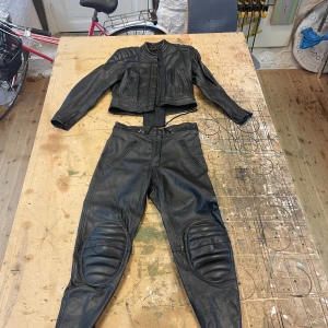 Svart skinnjacka med matchande byxor (motorcykel klädsel) - Säljer en svart skinnjacka med matchande byxor. Jackan har en tight passform med dragkedja och vadderade detaljer på axlar och armbågar. Byxorna har vadderade knän och dragkedja fram. I jackan sitter ett rygg skydd och det går att sätta ihop jackan med byxorna med blixtlås. Perfekt för motorcykelentusiaster!