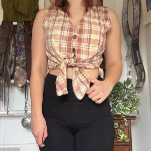 Rutig ärmlös blus med knyt - Köpt secondhand och knappt använd. Säljer en rutig ärmlös blus i beige och brun med knappar framtill och knyt i midjan. Perfekt för en avslappnad stil.