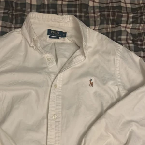 Vit skjorta från Ralph Lauren - Säljer en klassisk vit skjorta från Ralph Lauren med broderad logga på bröstet. Skjortan har en slim passform och långa ärmar med knappar framtill. Perfekt för en stilren look.